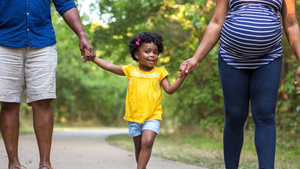 Child walking outdoors holding hand of adult on each side. Courtesy DEOHS.