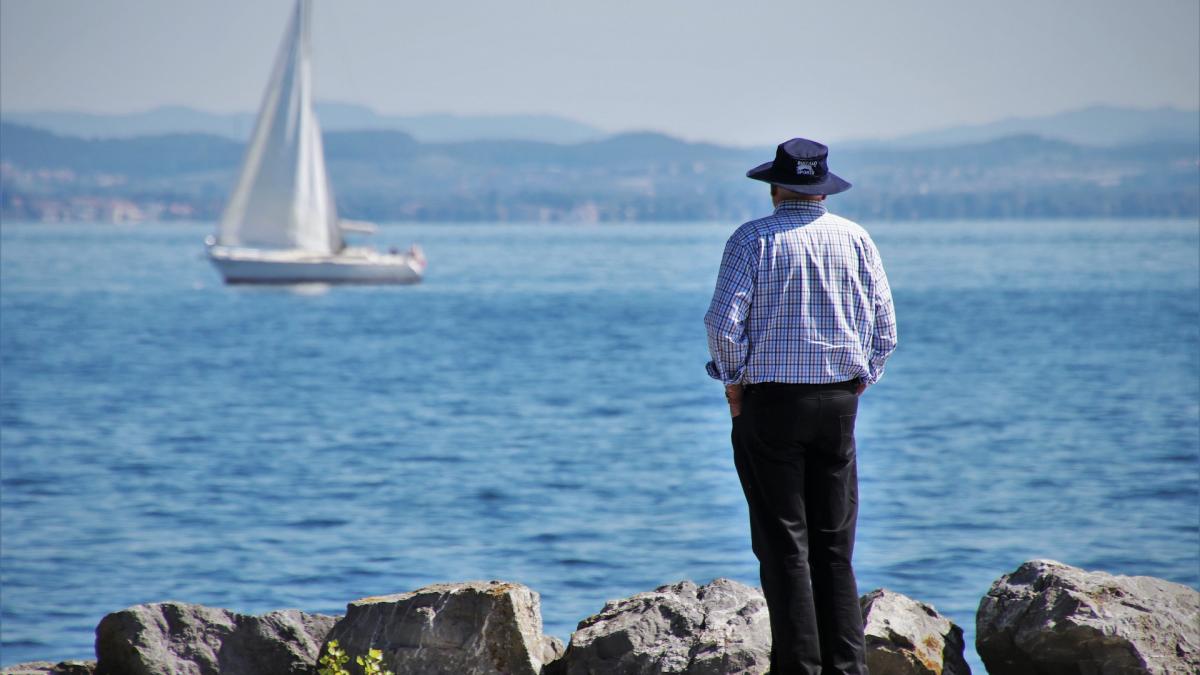 Man standing on shore looking out at a sailboat on the water