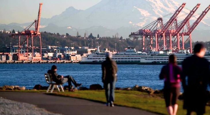 Photo of active people walking by the Seattle waterfront with ferry and mountain range prominently displayed in the background
