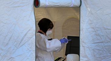 Healthcare worker standing inside door of tent wearing mask and protective gear and looking down at clipboard in hands