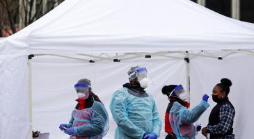 Masked and gowned health workers conduct COVID test in outdoor tent.