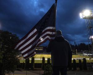 Photo taken behind a man holding an American flag looking down at a lit sportsfield at night